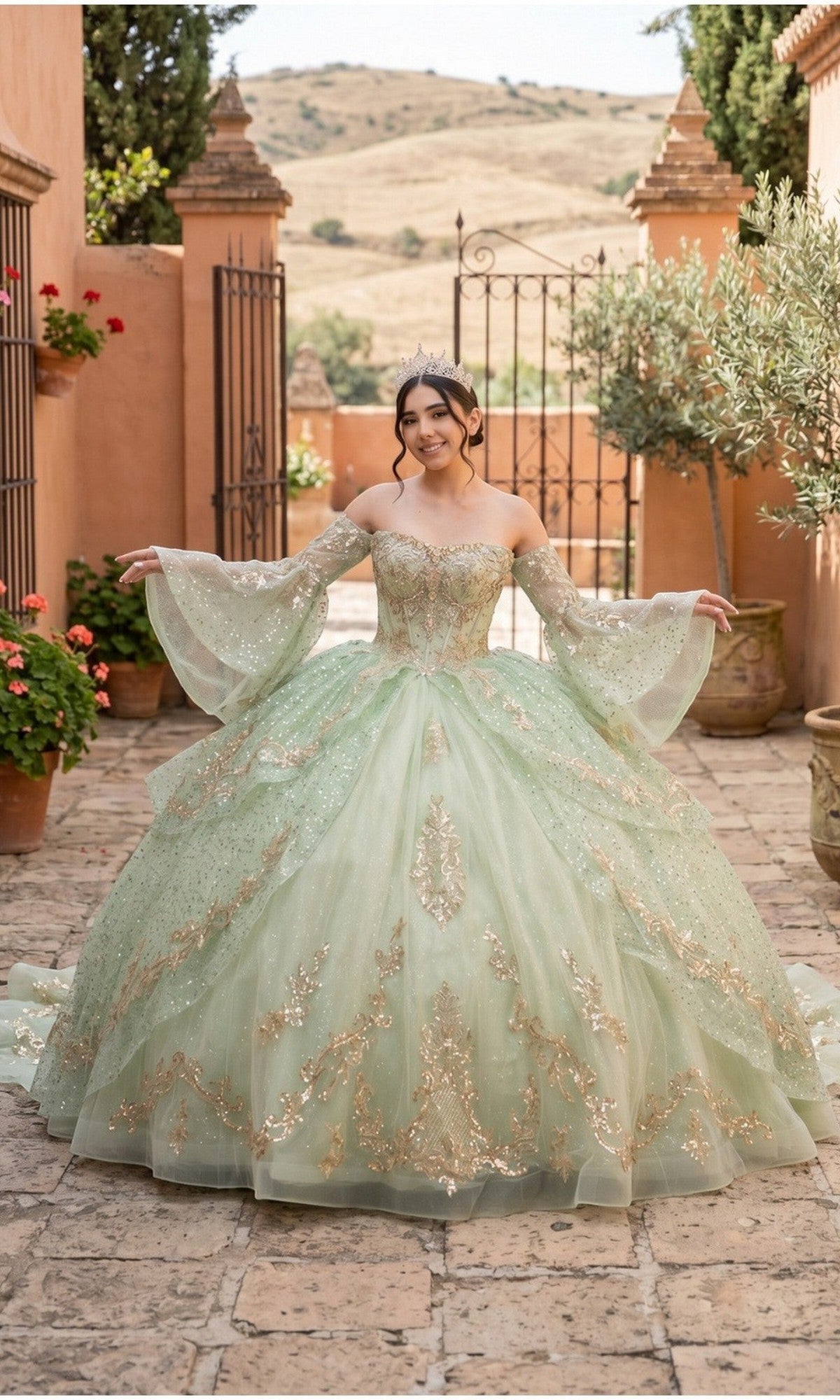 A young woman stands outside in a courtyard, smiling with arms extended, wearing the Quinceanera Dress 8575JQ by Cinderella Couture in sage. The gown features gold embellishments and long sheer sleeves, and its glitter tulle catches the light beautifully.